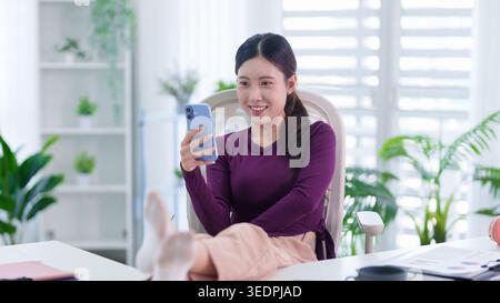 Asiatische Frau, die Videoanrufe auf dem Smartphone genießt, während sie vom Home Office aus in einem bequemen Stuhl mit Pflanzen sitzt Stockfoto
