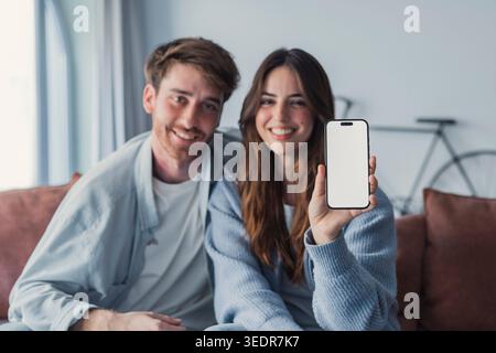 Ein glückliches Paar sitzt zu Hause auf einem Sofa und hält ein Smartphone mit einem leeren weißen Bildschirm, lächelt in die Kamera, repräsentiert Online-Shopping, digitale Dienste und Internetversicherung Stockfoto