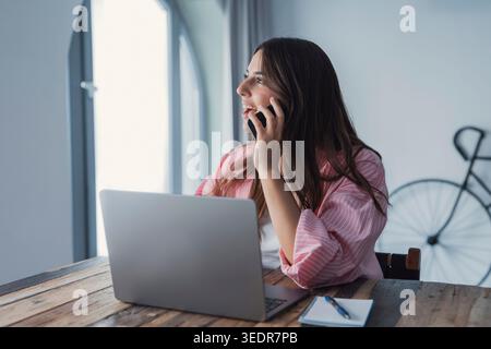 Eine junge professionelle Frau lächelt, während sie eine Kundin am Telefon an ihrem Schreibtisch berät, von ihrem Computer abgelenkt, aber ein freundliches und professionelles Verhalten aufrecht erhält Stockfoto