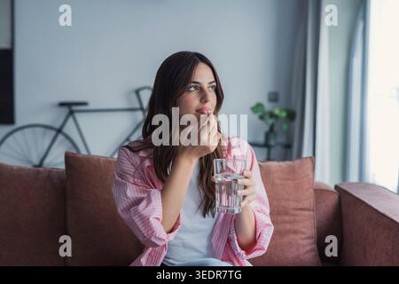 Eine glückliche junge Frau hält ein Glas Wasser und eine Pille zu Hause, genießt einen Moment der Selbstpflege und erhält ihre Gesundheit und ihr Wohlbefinden in einer positiven häuslichen Umgebung Stockfoto
