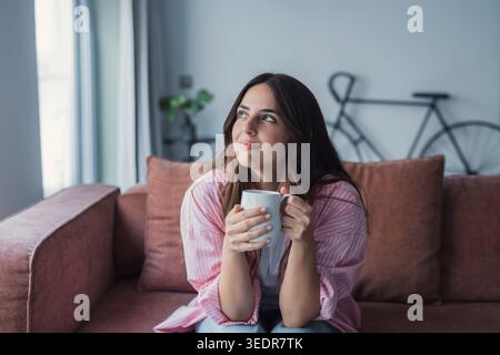 Eine glückliche junge Frau genießt einen ruhigen Morgen auf einem bequemen Sofa, hält ein heißes Getränk in der Hand, fühlt sich entspannt und stellt sich ihren anstehenden Arbeitstag und zukünftige Möglichkeiten vor Stockfoto