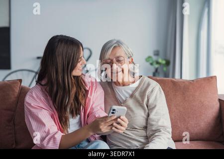 Lächelnde ältere Mutter und Erwachsene Tochter, die gemeinsam das Telefon benutzen, zu Hause auf einem gemütlichen Sofa sitzen, glückliche junge Frau und reife Mutter auf den Smartphone-Bildschirm schauen, Videos ansehen, Spaß haben, zwei Generationen Stockfoto