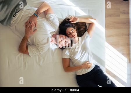 Liebevolles junges Paar, das zu Hause im Bett liegt, lächelt und umschließt, während es einen zärtlichen Moment teilt, Zuneigung, Nähe und Wärme während eines ruhigen, langsamen Wochenendes zum Ausdruck bringt Stockfoto