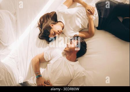 Ein glückliches junges Paar entspannt sich im Bett, lächelt und schaut sich an, während es einen zärtlichen Moment in gemütlicher Atmosphäre während eines ruhigen und faulen Wochenendes genießt Stockfoto