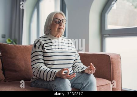 Achtsame gesunde reife Frau, die zu Hause Meditation praktiziert, ruhige ältere Dame mittleren Alters, die auf der Couch in Lotusstellung sitzt und Yoga für das geistige Gleichgewicht macht Luft atmen entspannend am stressfreien Wochenende Stockfoto