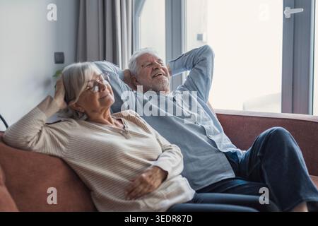 Ein grauhaariger Ehemann und eine Frau sitzen Seite an Seite auf einem Sofa mit Händen hinter dem Kopf, träumen zusammen und genießen Entspannung und emotionale Balance zu Hause Stockfoto