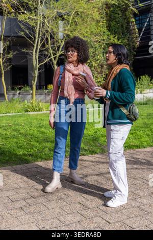 Verschiedene Freundinnen unterhalten sich, während sie Tassen zum Mitnehmen und ein Smartphone auf dem Weg zum Campus halten. Outdoor, Universität, Studenten, Lifestyle, Sonnenlicht, Stockfoto