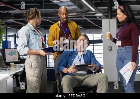 Verschiedene Kollegen im Büro um einen Mann mittleren Alters im Rollstuhl, der ein Tablet hält, die Karte teilen. Zusammenarbeit, Teamarbeit, offener Plan, Tageslicht, modern, c Stockfoto