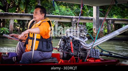 Thailändischer Langboot-Fahrer in einer orangen Schwimmweste fährt an einer Kanalwand am schwimmenden Markt Damnoen Saduak vorbei Stockfoto