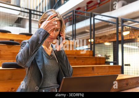 Professionelle Frau im Blazer, die am Telefon telefoniert, während sie im Coworking-Bereich mit einem Laptop arbeitet Stockfoto