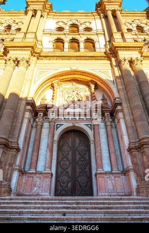Flacher Blick auf die kunstvoll verzierte Steinfassade und den geschnitzten hölzernen Haupteingang der Kathedrale von Malaga in Malaga, Spanien, beleuchtet durch Sonnenlicht gegen eine Clea Stockfoto