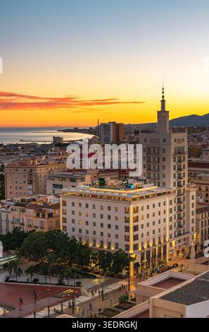 Blick auf Malaga, Spanien bei Sonnenuntergang, mit beleuchteten mittelhohen Gebäuden, einem markanten Turm, einer Dachterrasse und einem Stadtplatz mit Baum Stockfoto