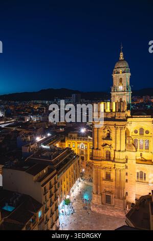 Blick auf den beleuchteten Turm der Kathedrale von Malaga, der sich über den umliegenden Gebäuden und dem plaza erhebt, mit weit entfernten Bergen unter einem Stockfoto