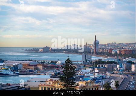Erhöhter Blick auf den Hafen von Malaga in Spanien mit Schiffen, Lagerhäusern, Lagertanks und Kränen entlang der Uferpromenade mit Wohnblöcken und der Skyline der Stadt Stockfoto