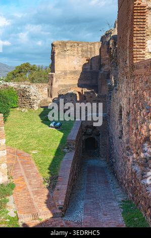 Alte Stein- und Ziegelfestungsruinen mit einem schmalen gemauerten Gang, der zu einem dunklen Tunnel führt, neben einem grasbewachsenen Innenhof unter einem teilweise bewölkten Himmel in Ma Stockfoto
