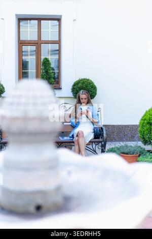 Eine Frau sitzt leise auf einer Bank im Garten. Sie konzentriert sich auf ihr Handy. Die Umgebung umfasst Grün und einen Brunnen. Es ist Tageslicht, erschaffen Stockfoto