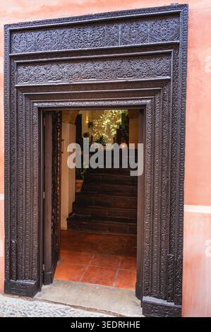 Vertikaler Blick auf eine verzierte dunkle Holztür mit komplizierten Schnitzereien, die sich auf Terrakottafliesen öffnen, und eine schmale Treppe, die nach oben führt, Deko Stockfoto