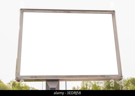 Eine leere Plakatwand befindet sich in einem von Bäumen umgebenen öffentlichen Raum. Der Himmel ist bewölkt und die Szene zeigt keine Werbung oder Nachrichten. Es wird angezeigt Stockfoto