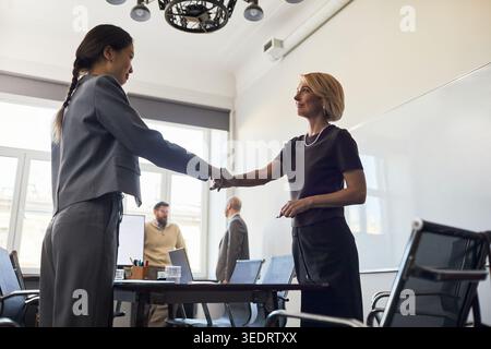 Weitwinkelaufnahme von zwei Geschäftsfrauen, die sich bei einem Büromeeting die Hand schütteln, um Partnerschaft und Verhandlungen zu signalisieren. Ideal für geschäftliche Teamarbeit oder Einstellungen Stockfoto