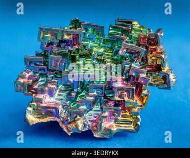 Schillerndes Wismut-Kristall mit faszinierenden geometrischen Trichterformationen, die in leuchtenden Regenbogenfarben im Metallic-Look gegen Blau glänzen. Stockfoto