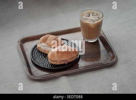 Auf einem Holztablett befindet sich ein strukturiertes Glas Eislatte neben einem dekorativen schwarzen Teller mit zwei Rollen, die mit herzhaftem Spinat und kombiniert sind Stockfoto