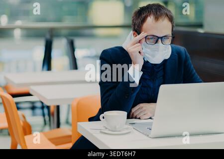Mann in medizinischer Maske und Brille, sitzt vor dem modernen Laptop-Computer, posiert in gemütlicher Cafeteria, liest Nachrichten über Coronavirus, besorgt über Gesundheit, trinkt Kaffee. Epidemische Quarantäne Stockfoto
