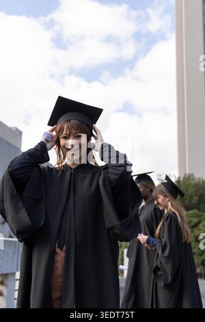 Andere Absolventen stehen auf dem Campus plaza, lächeln und passen schwarze Kleider und Mützen an. Beginn, im Freien, Terrasse, Turm, Bäume, Tageslicht, Bademantel Stockfoto