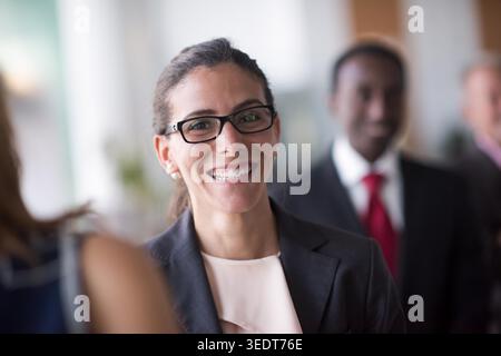 Lächelnde professionelle Frau mit Brille und Anzug, die in einer hellen Büroumgebung mit Kollegen interagiert und Vertrauen, Teamarbeit, Führungsstärke und positive Energie am Arbeitsplatz vermittelt. Stockfoto