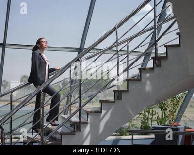 Selbstbewusste Geschäftsfrau in maßgeschneidertem Anzug klettert in einem modernen Firmenbüro eine Glas- und Stahltreppe und vermittelt Ehrgeiz, Führungsstärke und Vorwärtsdynamik für Start-up-, Karriere- und Berufskonzepte. Stockfoto