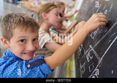 Ein lächelnder Grundschuljunge schreibt auf ein Schwarzes Brett, während Klassenkameraden zusammen zeichnen und lernen und Teamarbeit, Kreativität, Bildung und spielerische Konzentration in einem hellen Klassenzimmer vermitteln. Stockfoto