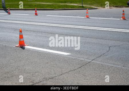 Verkehrskegel stehen in einer Reihe auf der Straße und markieren einen Abschnitt der Straße. Der Bürgersteig zeigt Anzeichen von Verschleiß, und die Sonne scheint hell über dem Boden. Stockfoto