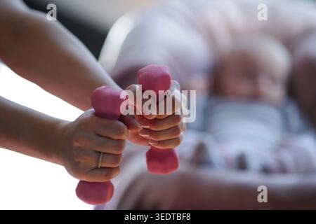 Eine Mutter hält rosa Kurzhanteln in den Händen. Ihr Baby liegt in einem Stuhl in der Nähe. Es ist hell und sie konzentriert sich auf ihr Workout. Stockfoto