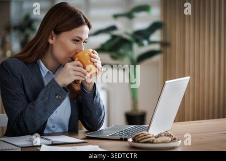Eine Frau im Anzug sitzt an einem Tisch in ihrem Haus und trinkt Kaffee aus einer orangen Tasse, während sie an ihrem Laptop arbeitet. Kekse werden auf dem Tisch in der Nähe platziert. Stockfoto