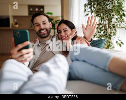 Ein Mann und eine Frau sitzen zusammen auf einer Couch, schauen auf ein Telefon und lächeln. Sie winken jemandem beim Videoanruf zu und zeigen Freude. Stockfoto