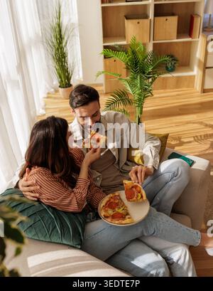 Ein Paar genießt Pizza auf einer Couch im Wohnzimmer. Sonnenlicht strömt durch das Fenster, während sie lächeln und gemeinsam Essen teilen. Stockfoto