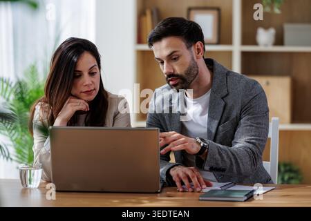 Zwei Kollegen diskutieren über die Arbeit, während sie an einem Schreibtisch sitzen. Vor ihnen ist ein Laptop offen. Sie scheinen in Gesprächen involviert zu sein. Stockfoto