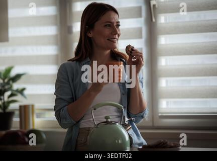 Eine Frau trinkt Tee und isst Kekse am Morgen. Sie steht in einer Küche mit Holzschränken Stockfoto