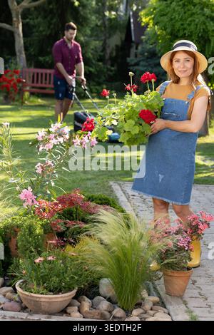 Ganzkörperporträt einer jungen Gärtnerin, die im Hinterhof steht und rote Blumen hält, während ihr männlicher Partner Gras im Hintergrund mäht Stockfoto