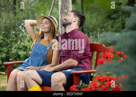 Mann und Frau entspannen auf einer Bank im Garten im Garten, während sie die Sommerbrise mit geschlossenen Augen genießen. Ein Paar ruht sich aus, nachdem es hart gearbeitet hat Stockfoto