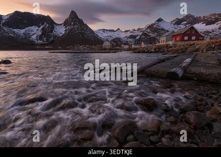 Unterirdische Meereslandschaft mit Wellen an einem felsigen Ufer und einem kleinen Küstendorf unter schneebedeckten Bergen. Stockfoto
