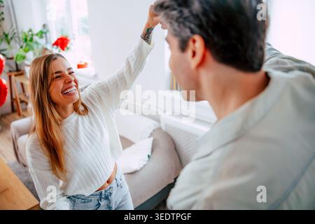 Glückliche junge Frau lächelt beim Tanzen mit Freund zu Hause, romantisches Paar genießt spielerische Momente in hellem, modernem Wohnzimmer Stockfoto