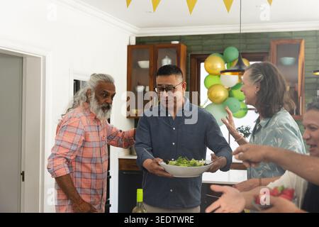 Ältere asiatische Männer und reife Frau plaudern in der Küche, halten Salatschüssel und Wein in der Nähe. Feiern, Sammeln, Ballons, Flaggen, Dekor, fest Stockfoto