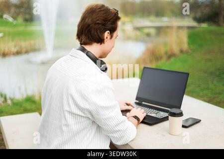 Mann, der an einem Laptop arbeitet, an einem Tisch im Freien in einem Park nahe einem Brunnen. Konzept der Fernarbeit, des Lebensstils digitaler Nomaden und der Freiberufung mit Kaffee und Stockfoto