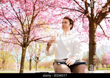 Ein lächelnder Mann sitzt auf einer Parkbank mit Laptop und Kaffee unter blühenden Kirschblüten. Fernarbeit, Freiberufung und Lebensstil im Frühling in Stockfoto