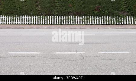 Weiß lackierter Holzzaun am Straßenrand mit hohem Zaun hinten. Bürgersteig und Straße vor dem Hotel. Hintergrund für Kopierbereich. Stockfoto