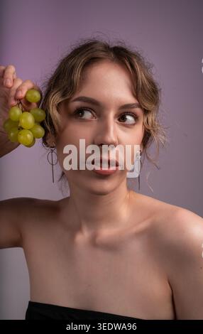 Junge Frau, die grüne Trauben hält, wegschaut, mit modernem Schmuck, posiert auf einem lila Hintergrund Stockfoto