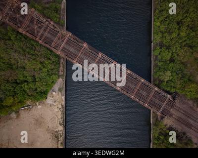 Aus der Vogelperspektive sehen Sie eine Eisenbahnbrücke aus rostigem Metall, die über einen dunklen Kanal führt, der von üppigen grünen Frühlingsbäumen in den Vororten von Chicago flankiert wird Stockfoto