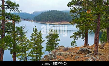 Pine Forest Lake Mit Misty Hills. Malerischer See, umgeben von Kiefern und felsigem Ufer, mit bewaldeten Hügeln und weichem Nebel im Hintergrund. Stockfoto