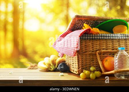 Picknickkorb mit Obst, Tellern und Wasserflasche auf einem Holztisch in einem sonnigen Park mit warmem Bokeh Hintergrund. Stockfoto