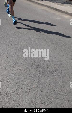 Läufer, die beim Outdoor-Training bei hellem Sonnenlicht lange Schatten auf der Asphaltstraße werfen, Fitnesskonzept Stockfoto